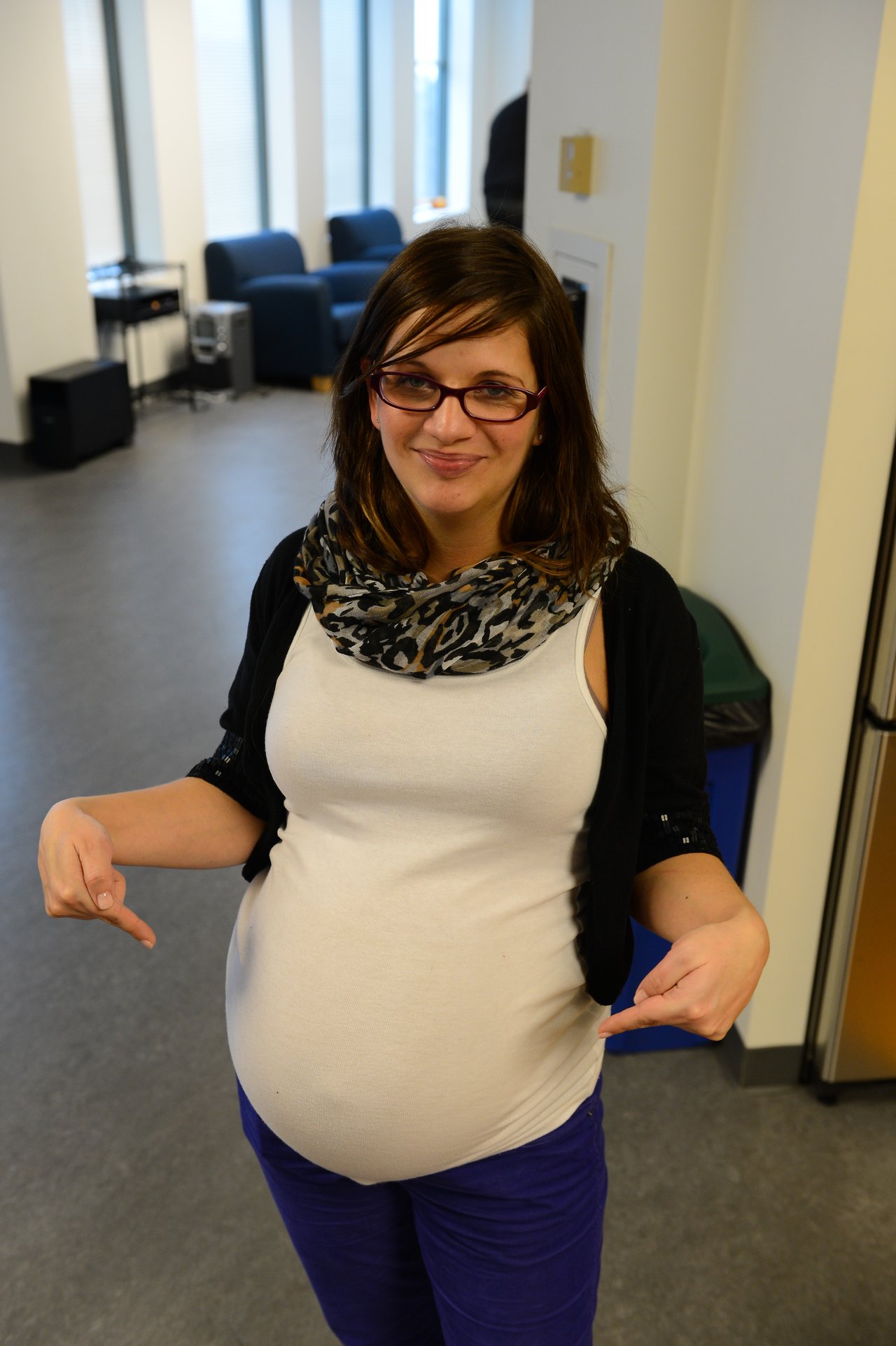 A smiling woman wearing glasses points to her pregnant belly while standing in an indoor setting.
