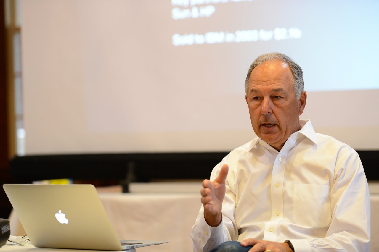 A man in a white shirt speaks while gesturing, with a laptop and presentation screen in the background.