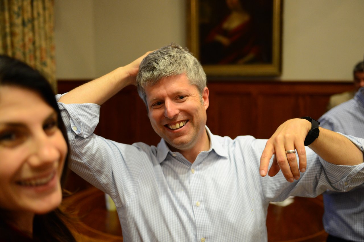A smiling man in a light blue shirt poses playfully, while a woman in the foreground also smiles.