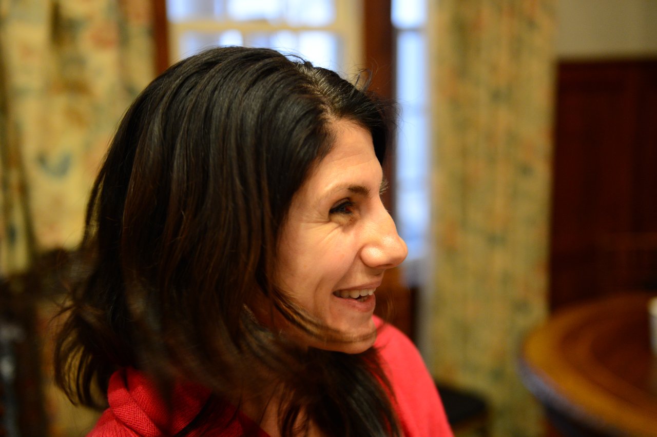 A woman with dark hair and a red top smiles while looking to the side in an indoor setting.
