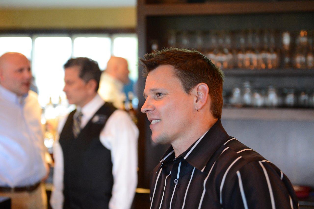 A person in a striped shirt smiles while standing in a bar or restaurant, with others conversing in the background.