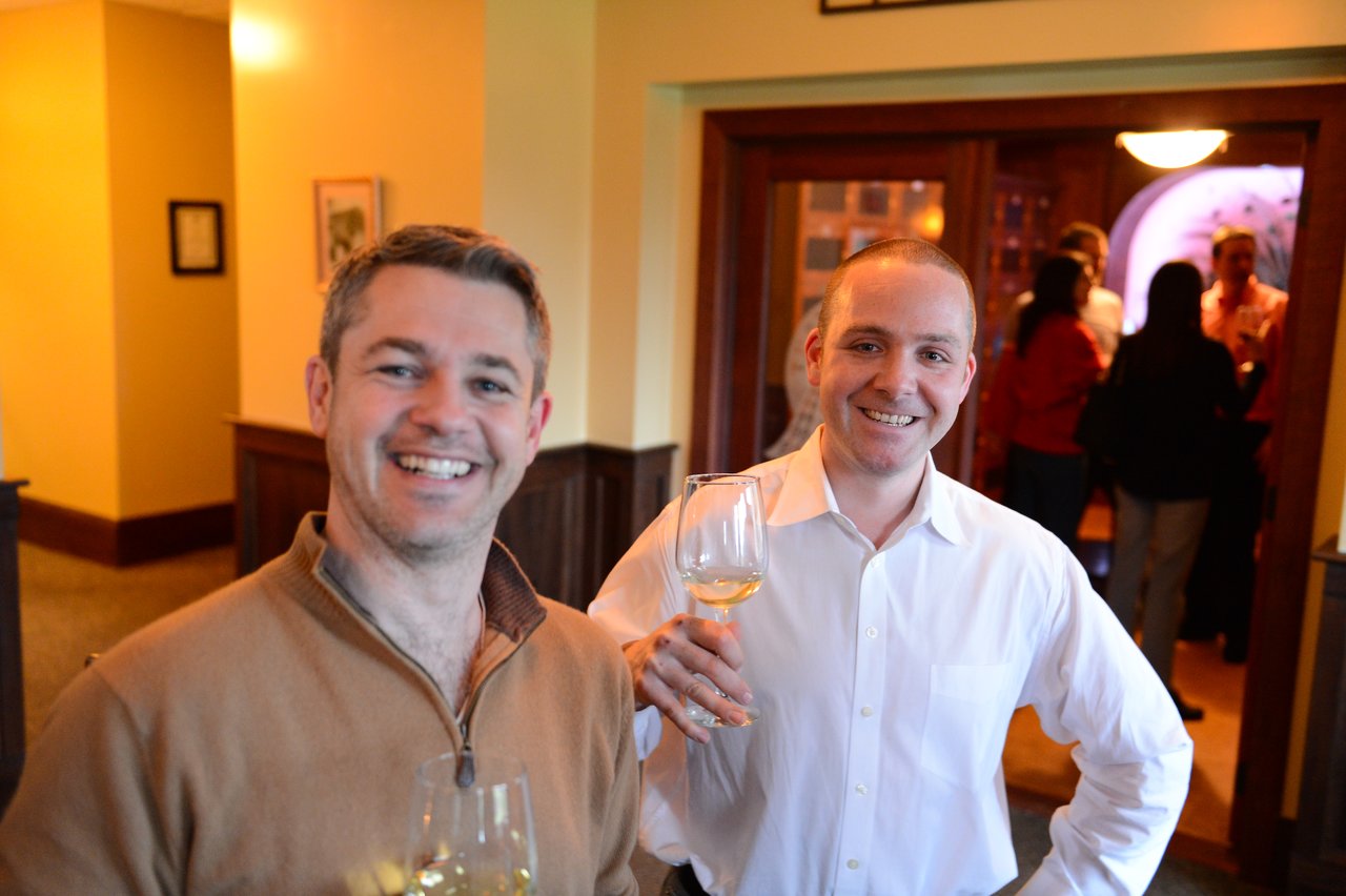 Two smiling men holding wine glasses at a social gathering, with a group of people conversing in the background.