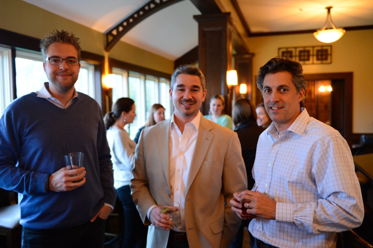 Three men stand together at an indoor gathering, holding drinks and smiling, while others converse in the background.