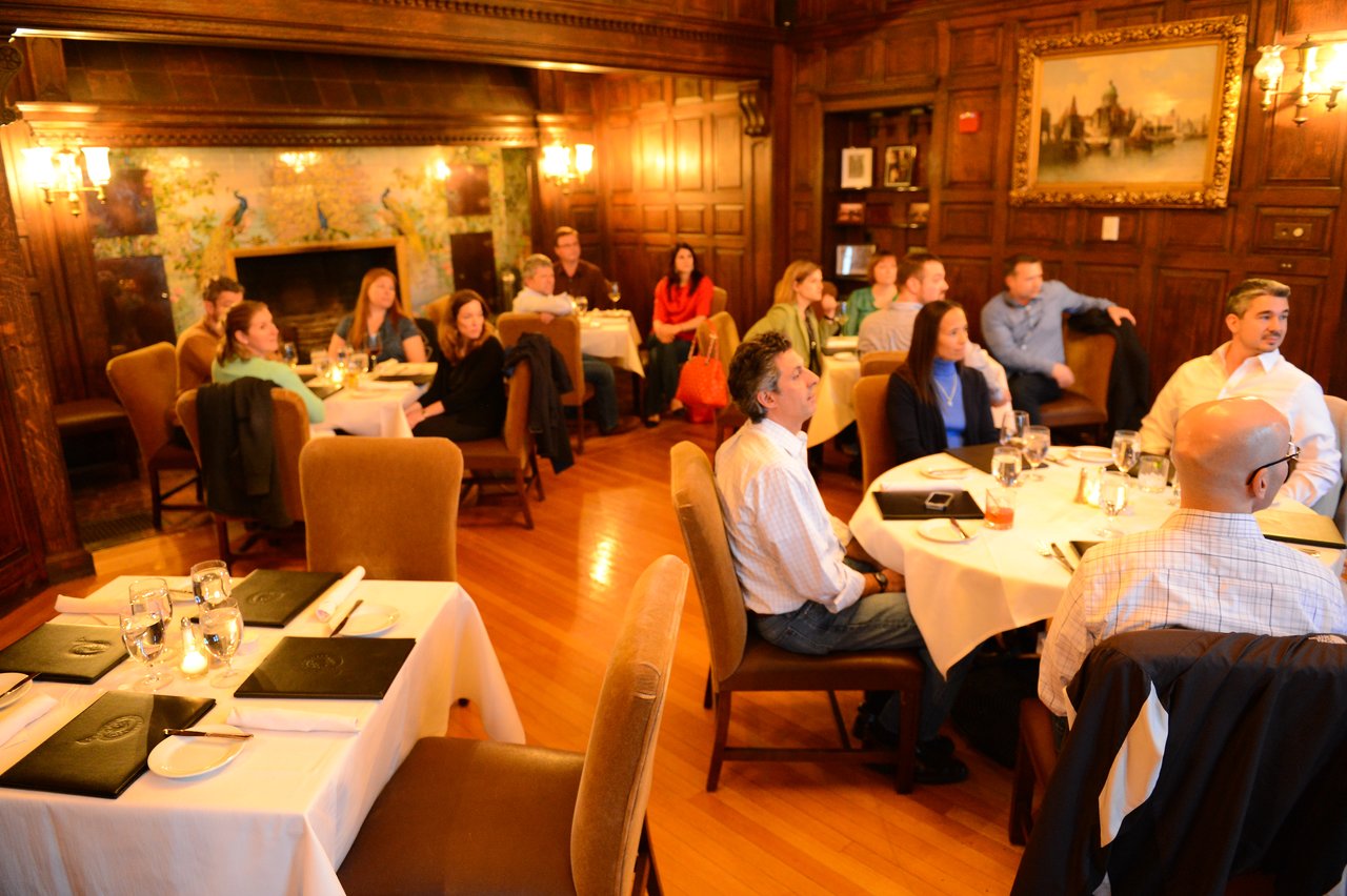 A group of people sits at round tables in a wood-paneled room, attentively listening to a presentation.