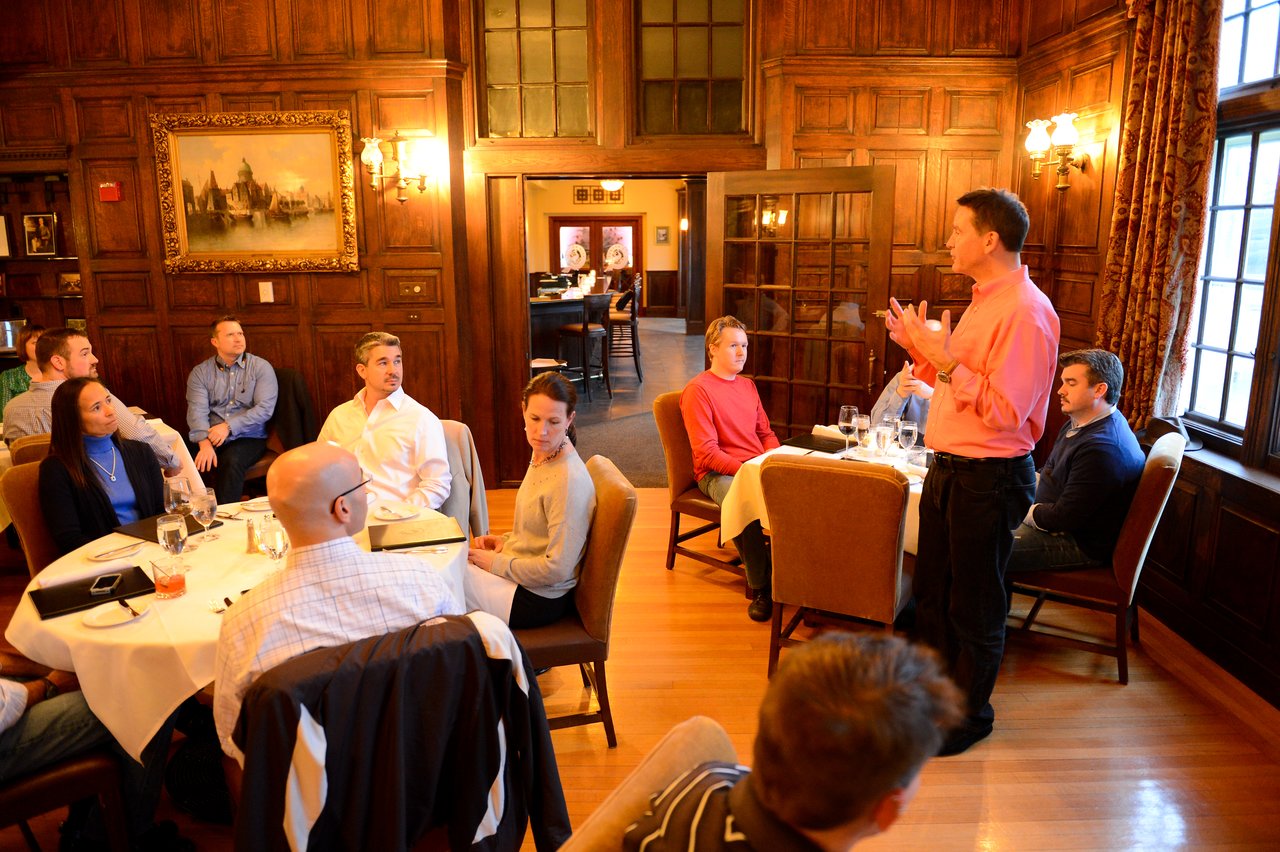 A man in a pink shirt stands and speaks to a group of people seated at tables in a restaurant.