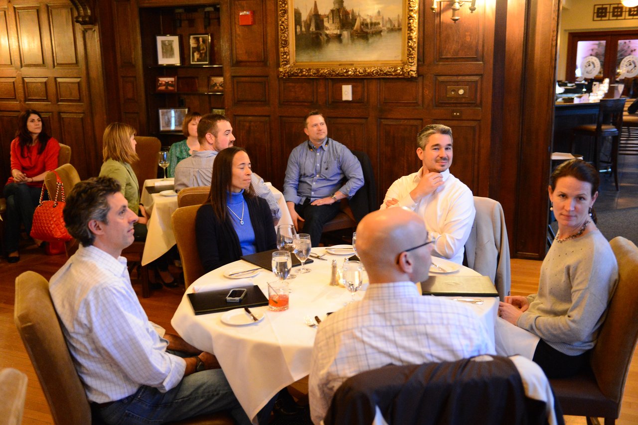 A group of people sits around a table in a restaurant, attentively listening to someone off-camera.