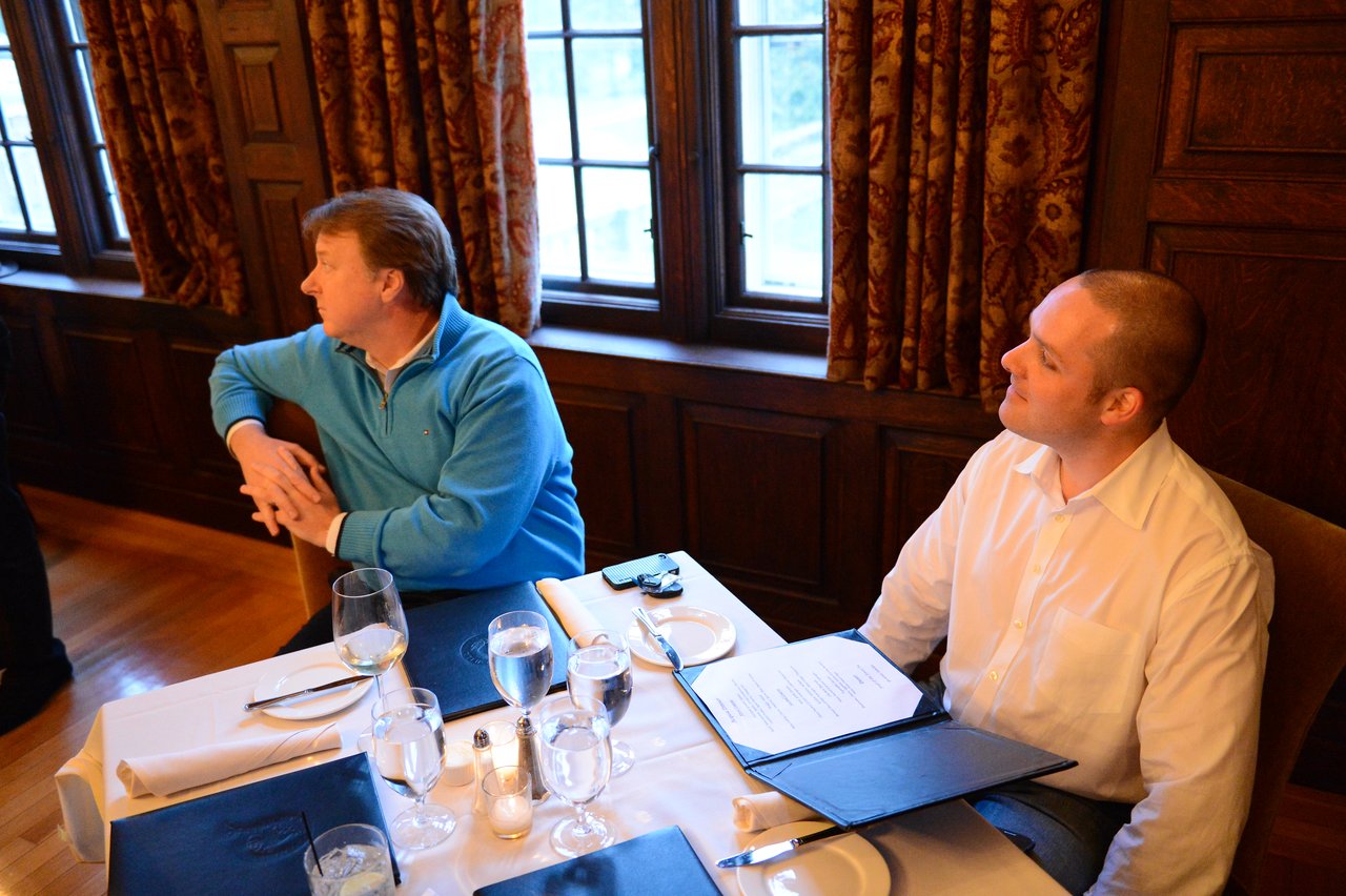 Two men sit at a restaurant table with menus and drinks, engaged in conversation and looking in different directions.