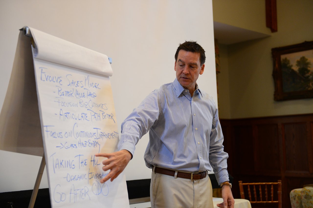 A man in a striped shirt gestures toward a flip chart with handwritten notes during a presentation.
