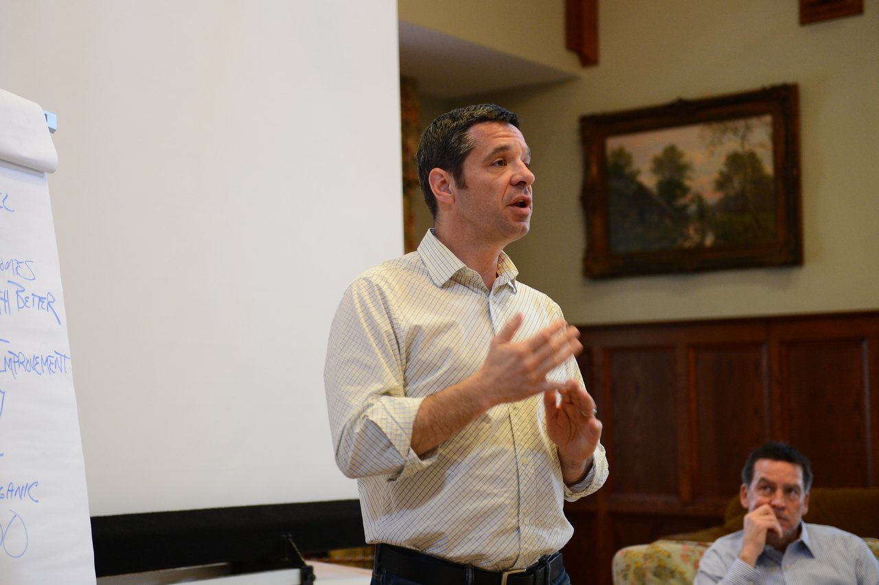 A man in a light-colored shirt gestures while speaking to a group, with a flip chart beside him.