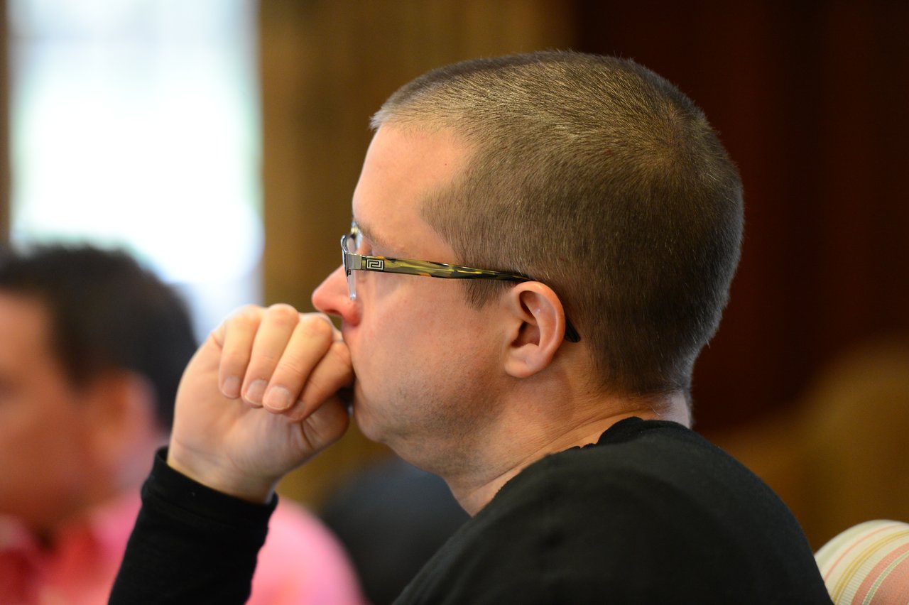 A person wearing glasses and a black shirt sits with their hand on their chin, appearing to listen attentively.
