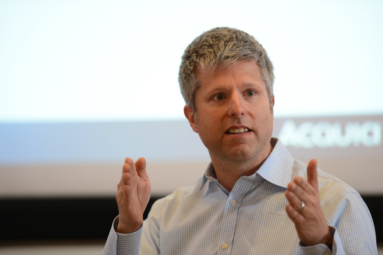 A person in a button-up shirt gestures with both hands while speaking in front of a presentation screen.