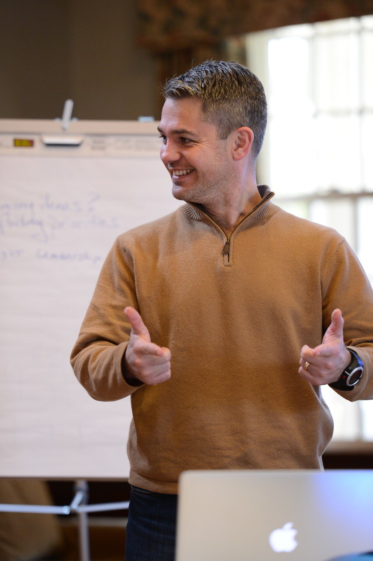 A man in a brown sweater gestures with both hands while speaking in front of a flip chart.