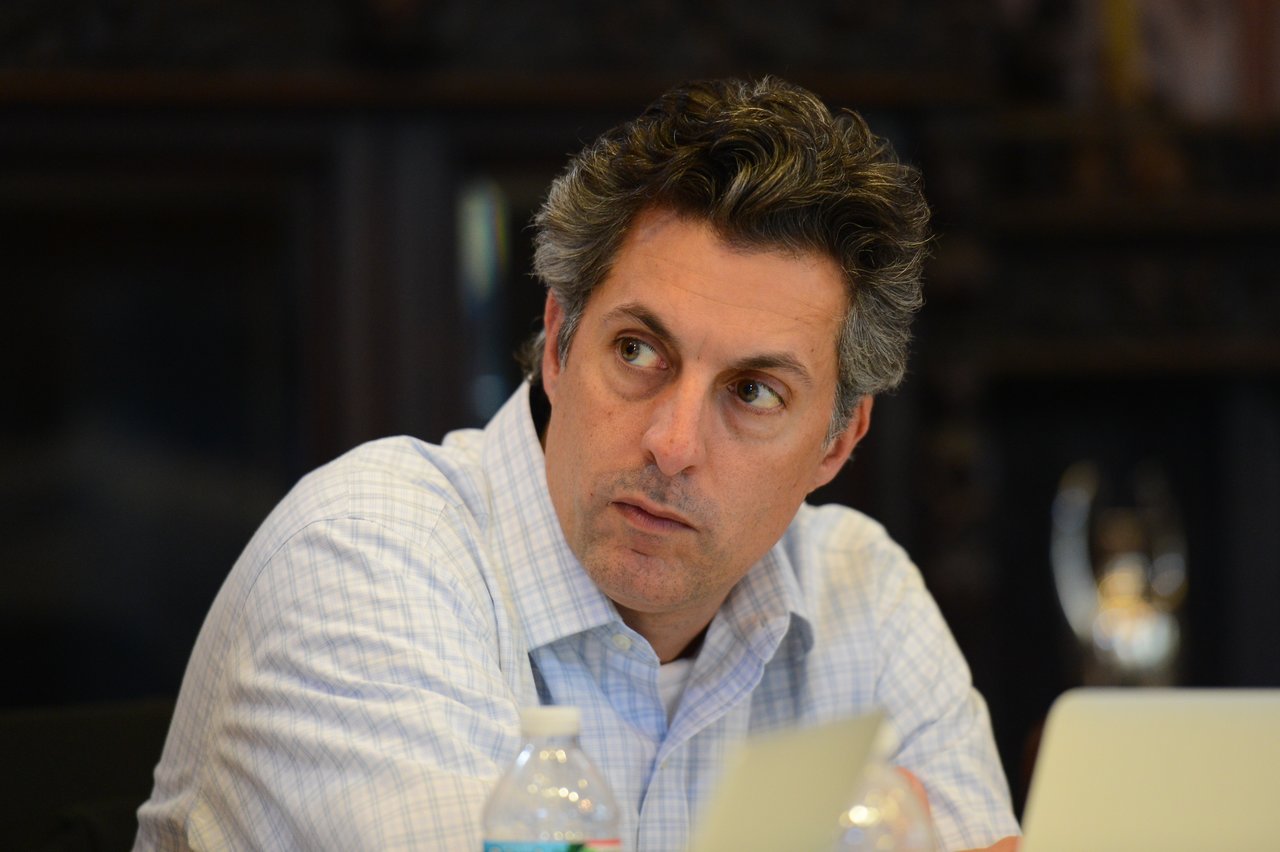 A man in a white shirt sits at a table, looking to the side with a focused expression.