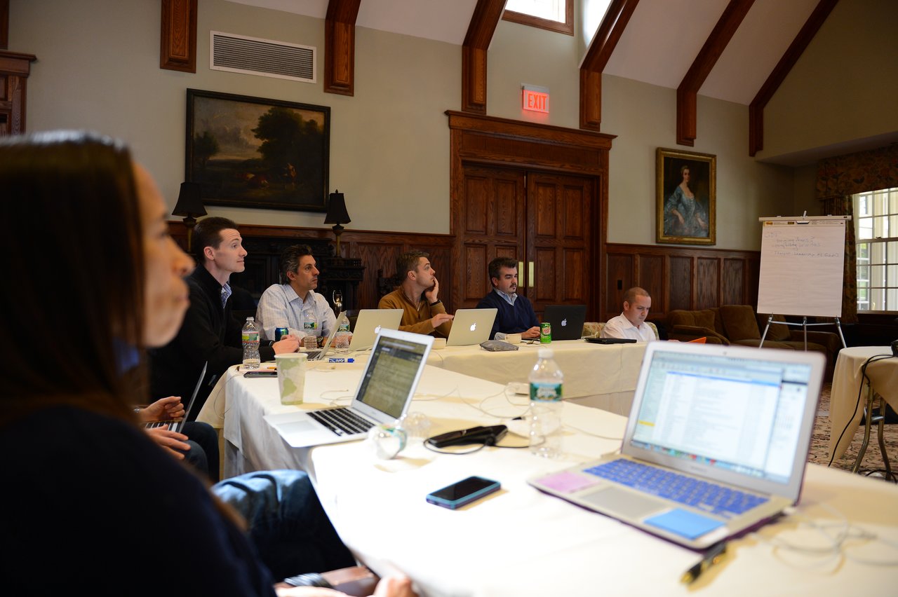 A group of people in a meeting room, working on laptops and listening to a discussion.