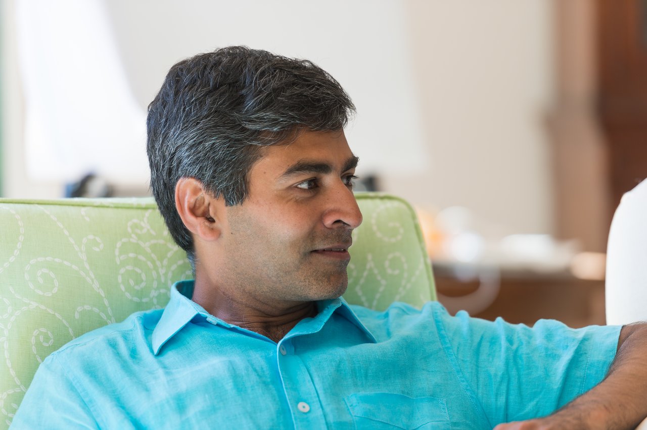 A man in a blue shirt sits on a green chair, looking to the side and engaged in conversation.