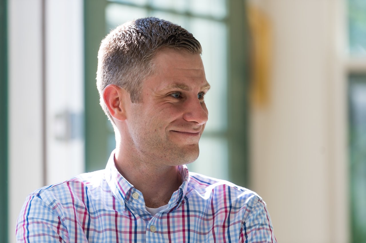 A man in a plaid shirt smiles and looks to the side in a well-lit indoor setting.