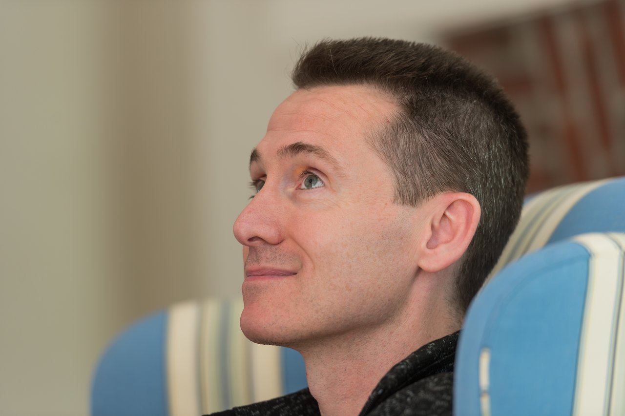A man sits in a striped chair, looking up with a slight smile.