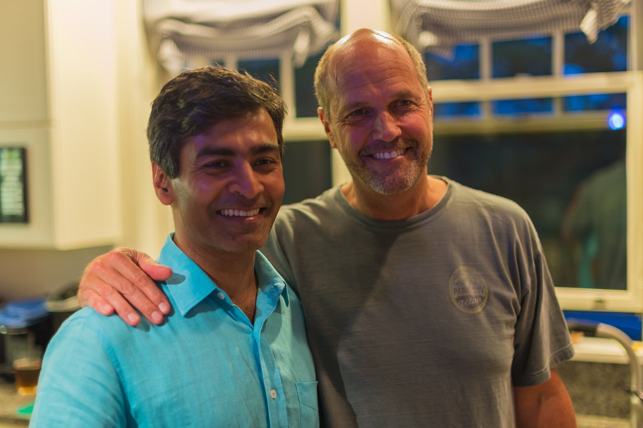 Two smiling men pose together indoors, one with his arm around the other.