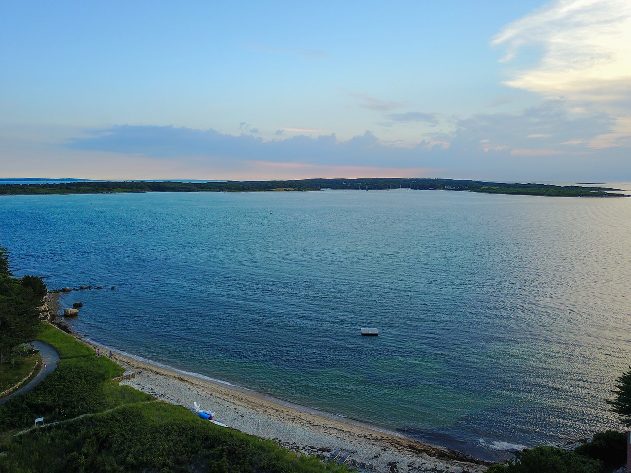 A calm coastal view with a sandy beach, blue water, a small floating dock, and distant land on the horizon.