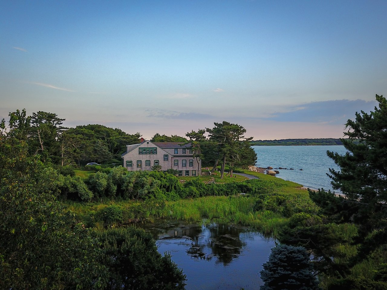 A large house surrounded by trees sits near the water, with a small pond in the foreground.