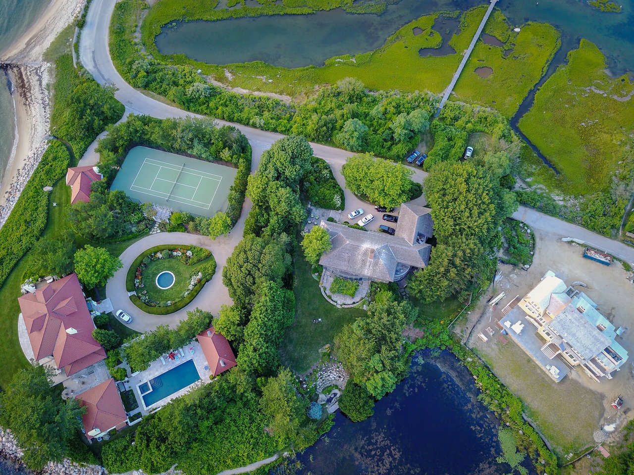 Aerial view of a coastal estate with houses, a tennis court, a swimming pool, and a pond surrounded by greenery.