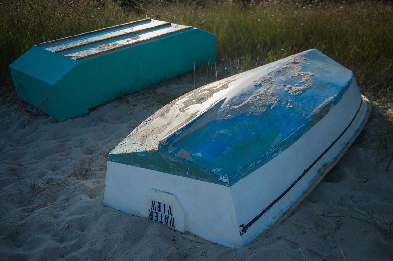 Two small boats are flipped upside down on a sandy shore, with grass in the background.