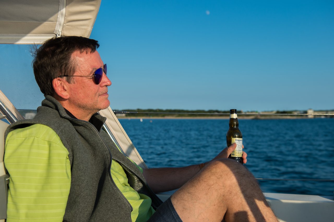 A man wearing sunglasses relaxes on a boat, holding a beer, with water and shoreline in the background.