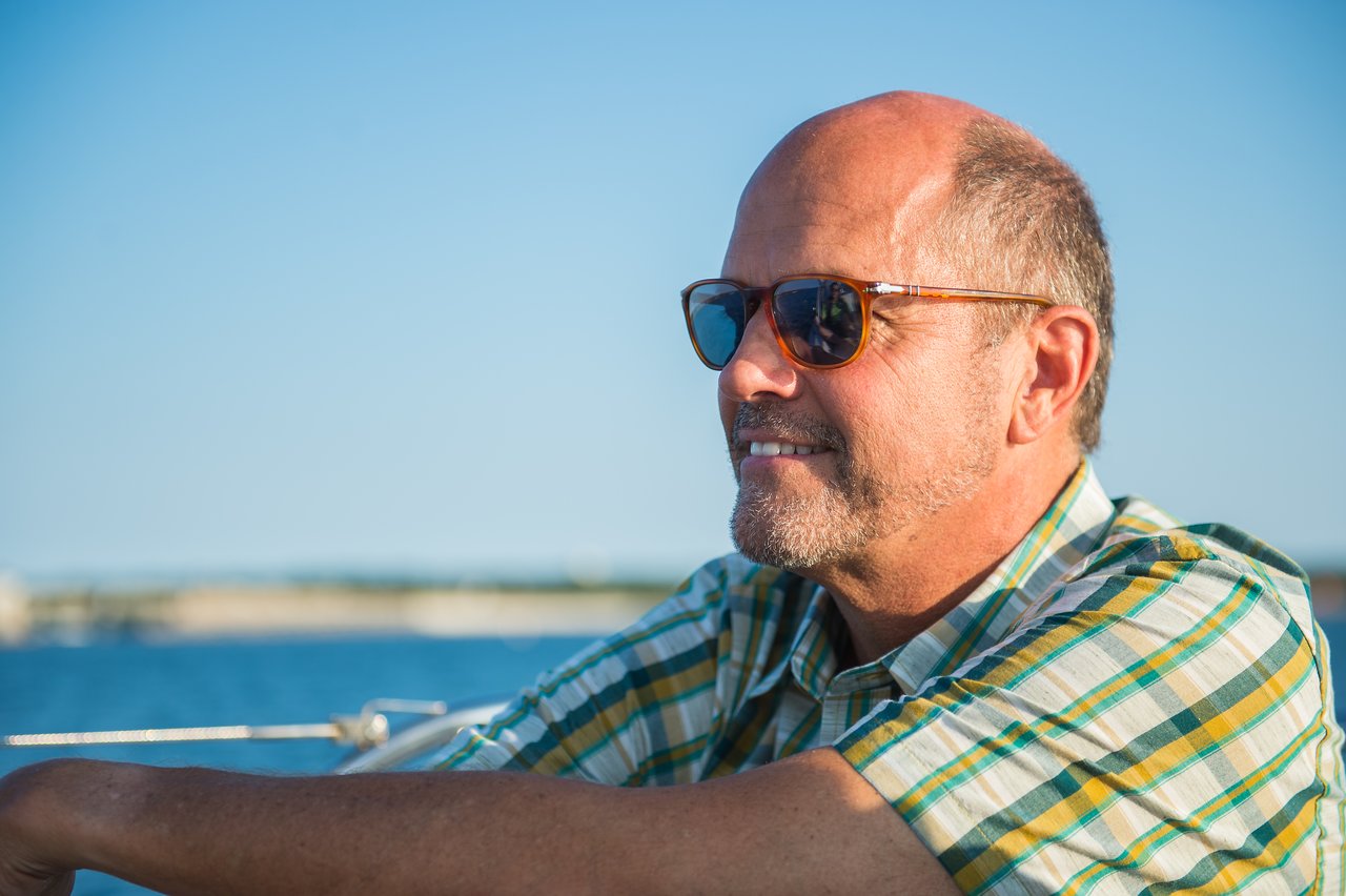 A man wearing sunglasses and a plaid shirt smiles while sailing on the water.