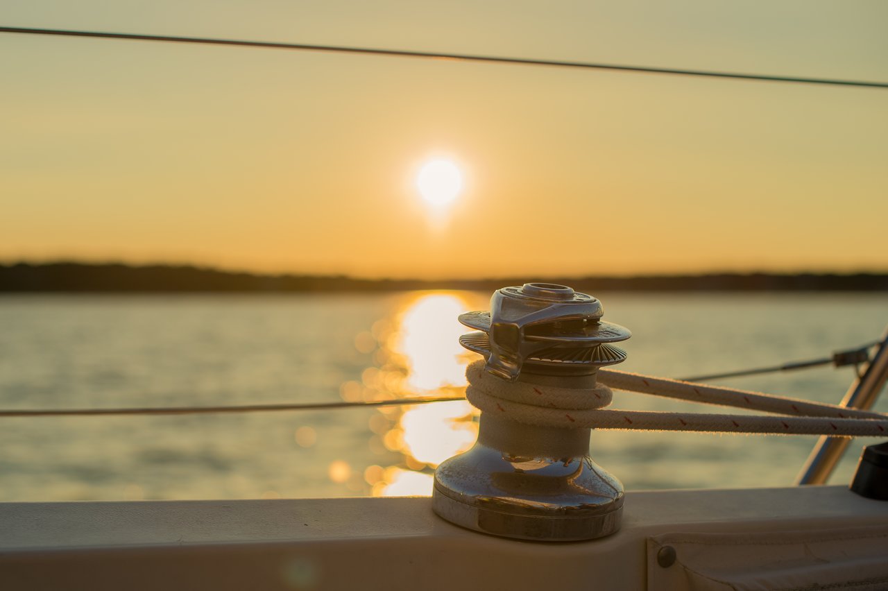 A sailboat winch with ropes in focus, with the sun setting over the water in the background.