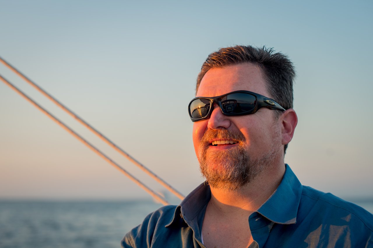 A man wearing sunglasses and a blue shirt smiles while sailing at Martha's Vineyard during sunset.