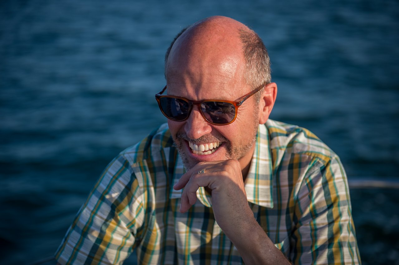 A man wearing sunglasses and a plaid shirt smiles while sailing on the water.