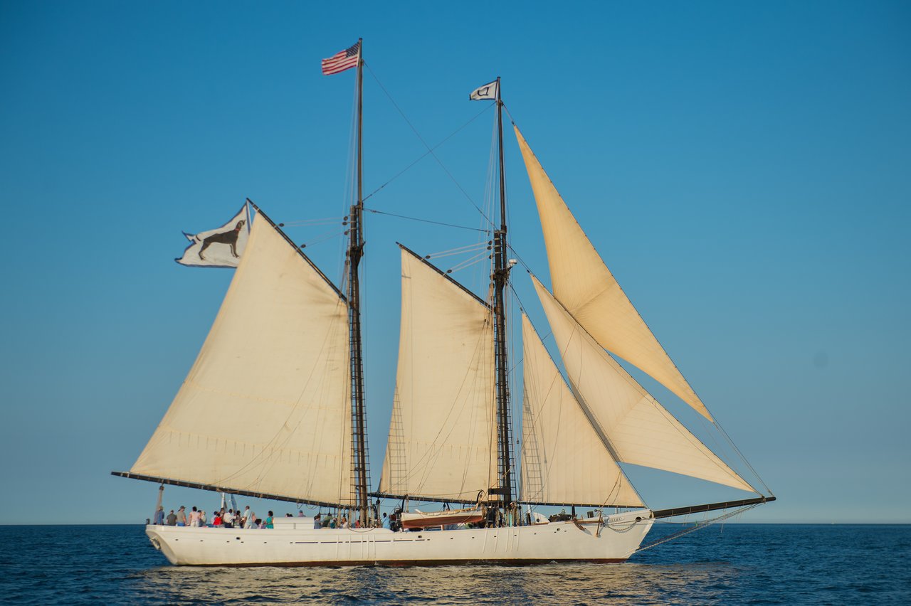 A large sailboat with multiple sails glides on the water, carrying a group of people under a clear sky.