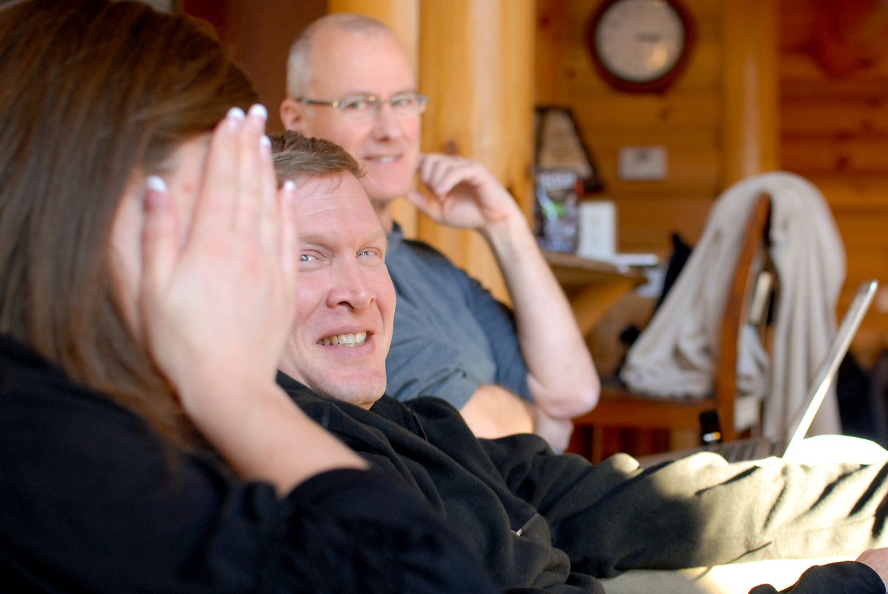 Three people sitting indoors; one person smiles at the camera while another covers their face with a hand.