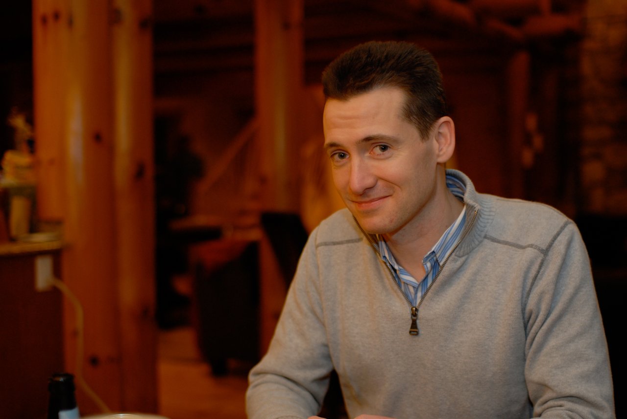 A man in a gray sweater sits at a table, smiling slightly and looking at the camera.