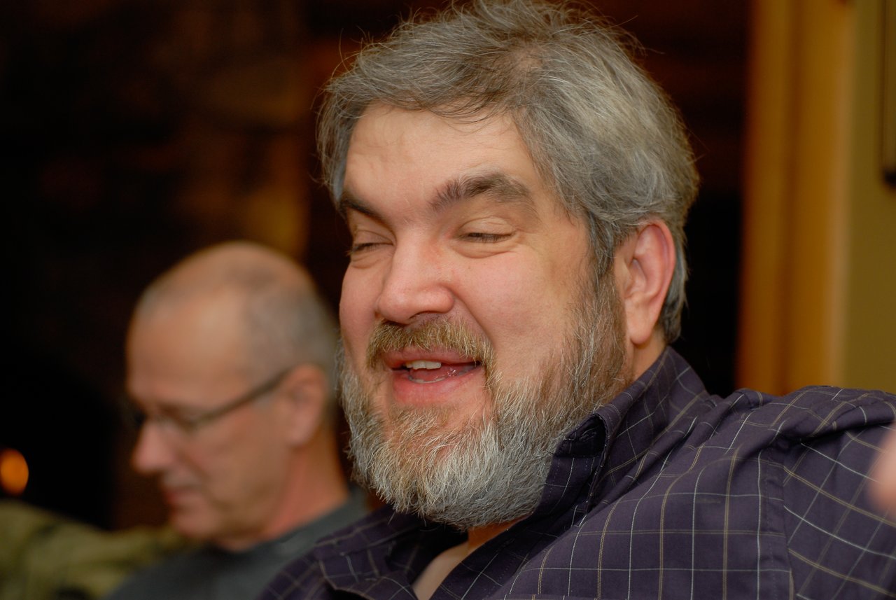 A bearded man in a checkered shirt smiles, while another man in glasses sits slightly out of focus behind him.