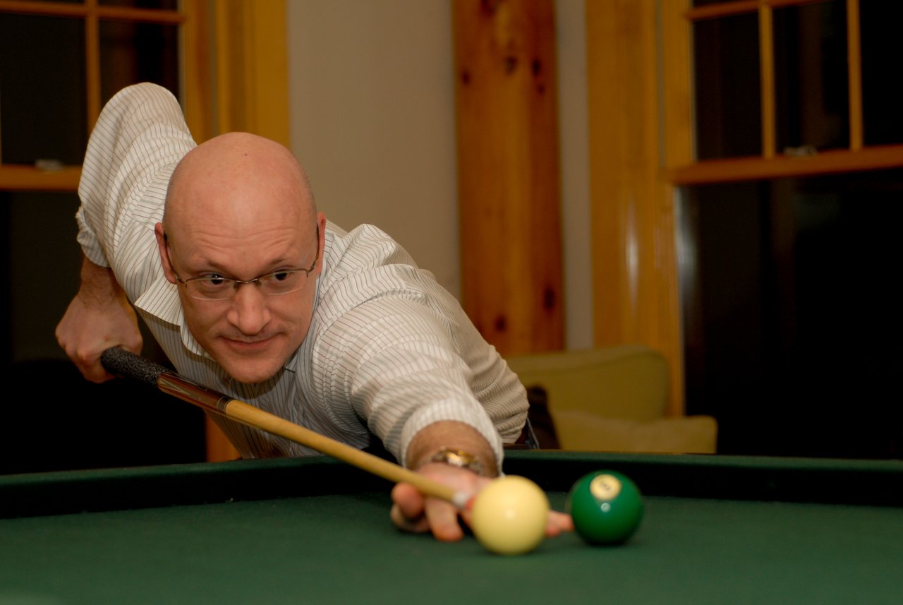 A man in glasses and a white shirt leans forward, aiming a pool cue at a ball on the table.