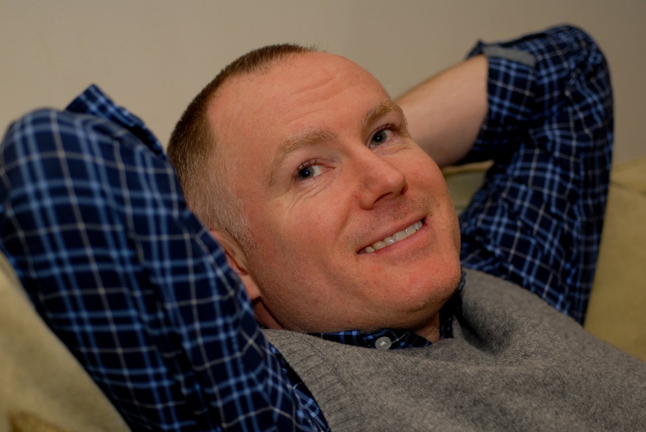 A man in a plaid shirt and vest leans back on a couch, smiling and relaxing.