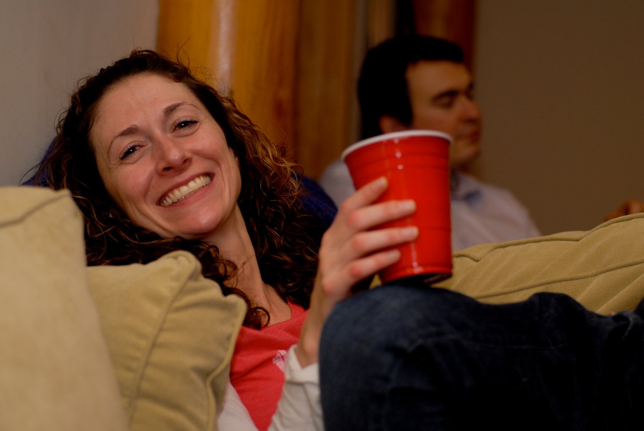 A smiling woman reclines on a couch, holding a red cup, while a man sits in the background.