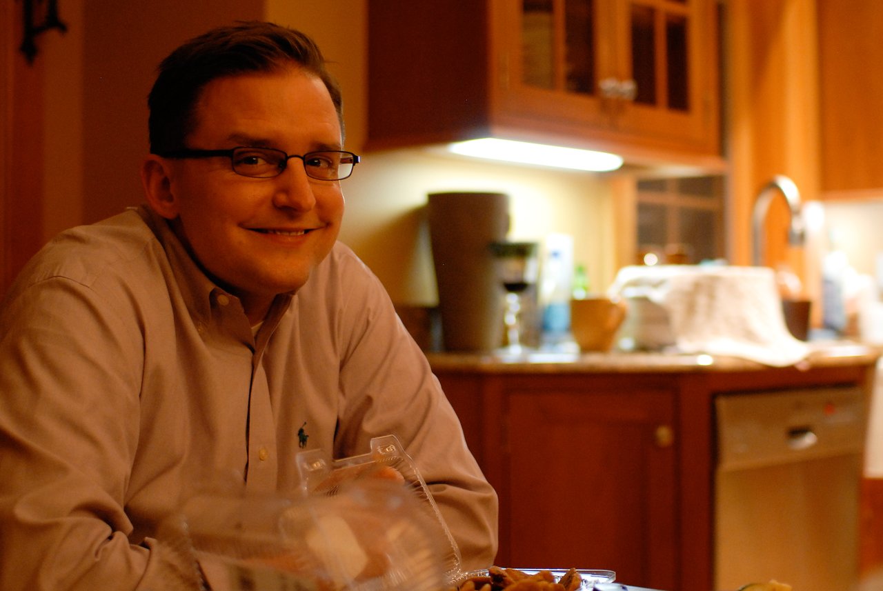 A man wearing glasses and a button-up shirt smiles while sitting at a table with food in a kitchen.