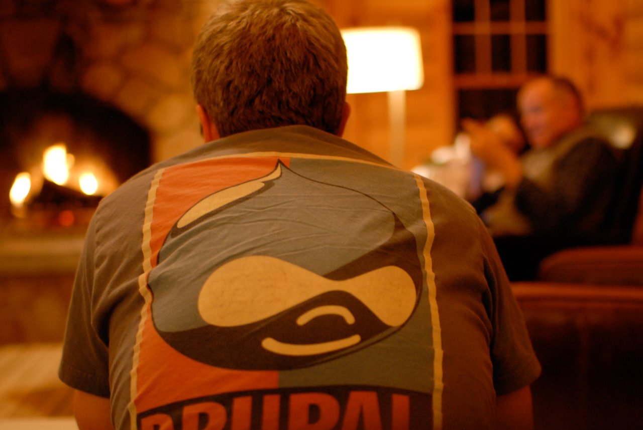 A person wearing a Drupal t-shirt sits by a fireplace, facing another person engaged in conversation.