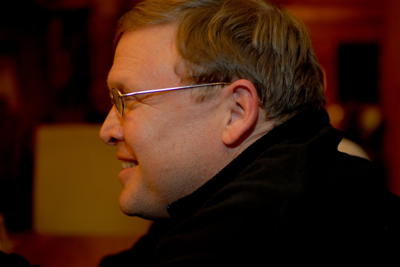 A man wearing glasses and a black jacket smiles while looking to the side in a dimly lit setting.