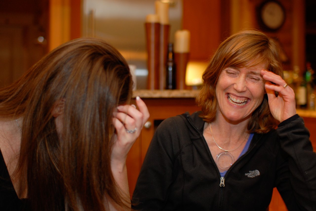 Two women laughing, one covering her face with her hand, in a warmly lit indoor setting.