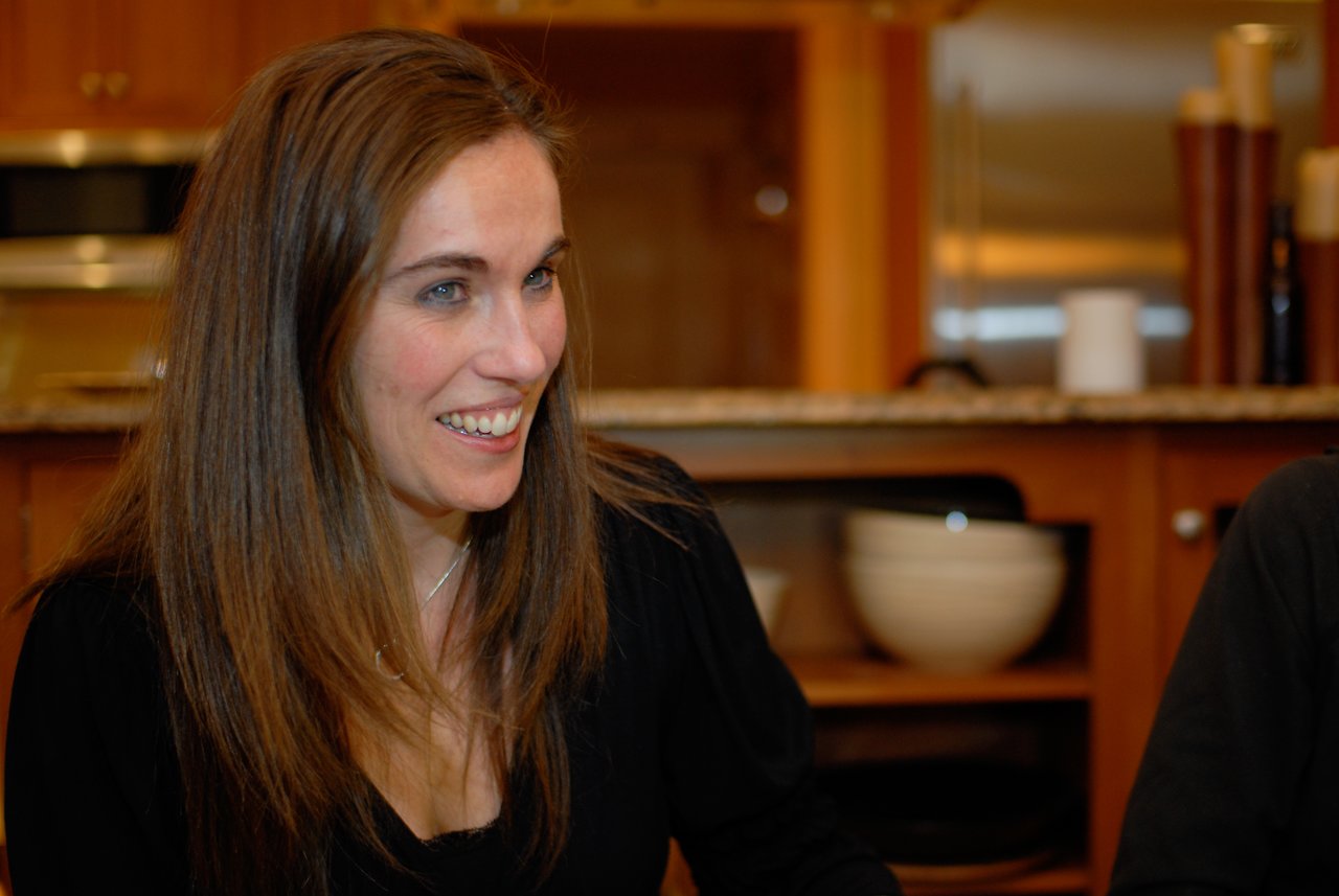A woman with long brown hair smiles while sitting indoors, engaged in conversation.
