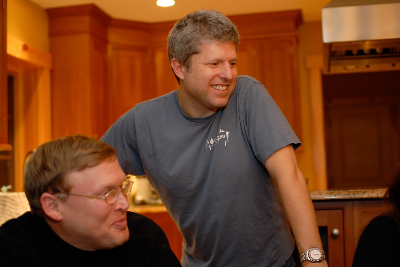 A man in an Acquia t-shirt leans on a counter, smiling, while another man sits nearby.