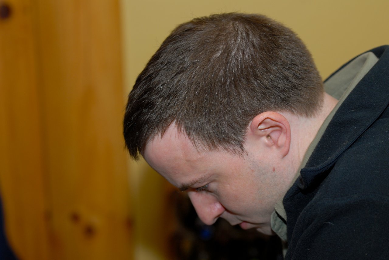 A man in a black jacket looks downward with a focused expression in an indoor setting.