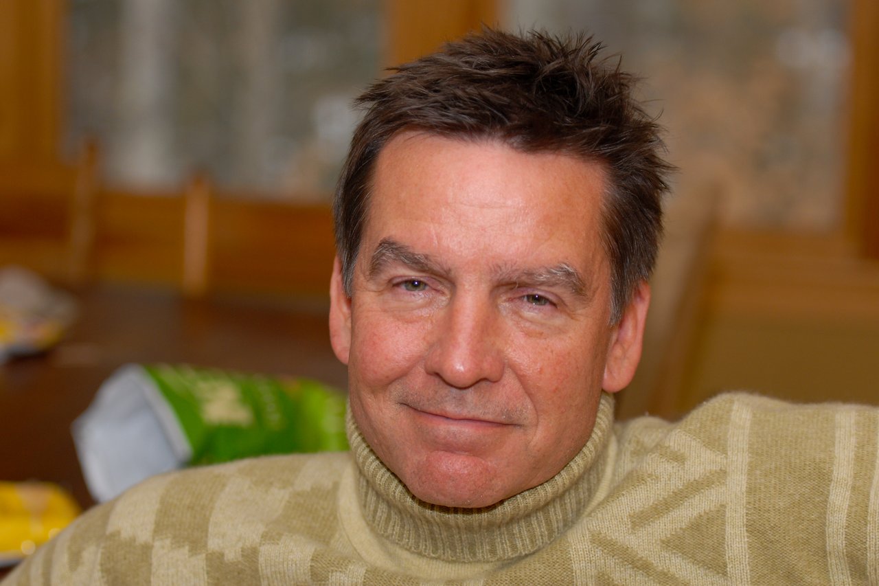 A man in a green turtleneck sweater sits indoors, looking directly at the camera with a relaxed expression.