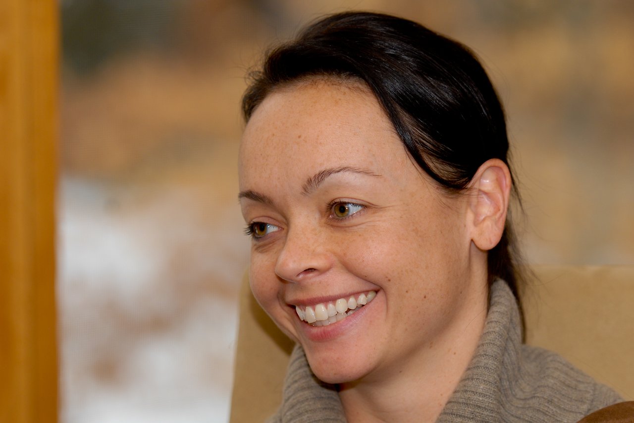 A woman with dark hair and a beige sweater smiles while looking slightly to the side.