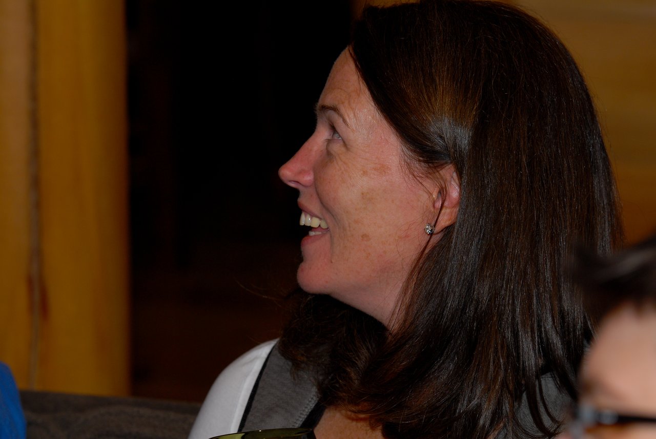 A woman with long brown hair smiles and looks to the side during an indoor event.