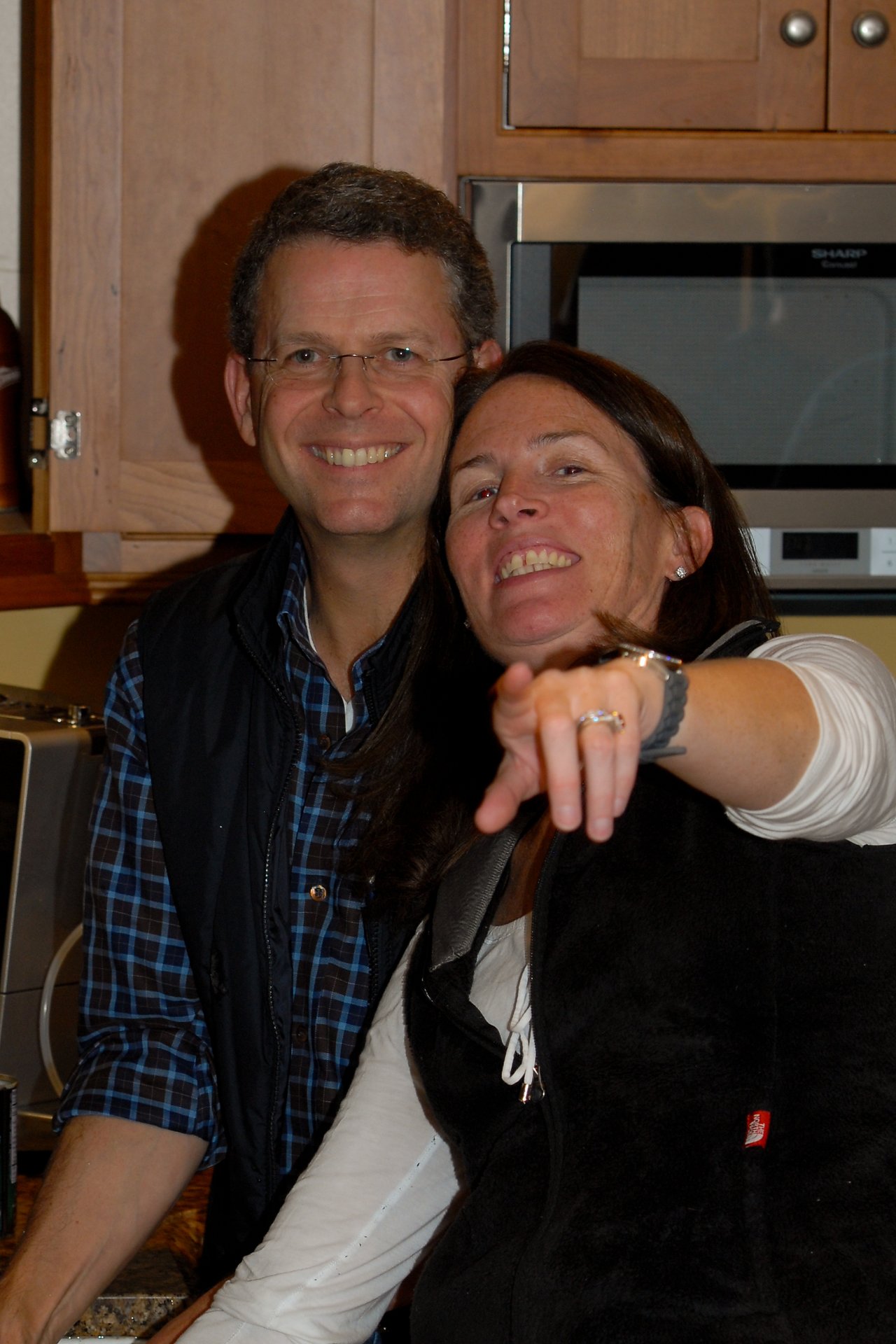 A smiling man and woman pose together in a kitchen, with the woman pointing towards the camera.