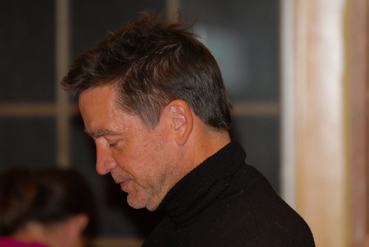 A man in a black turtleneck looks downward with a focused expression in an indoor setting.